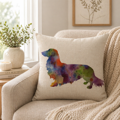 A square beige cushion cover pillow with a colourful watercolour dachshund silhouette, resting on a beige armchair in a cosy living room setting.