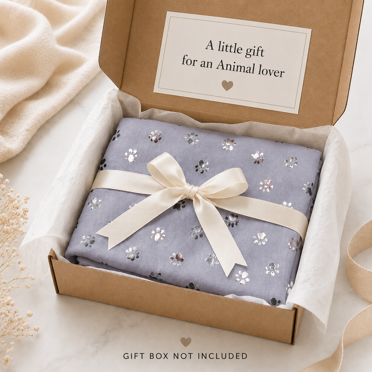A folded grey scarf with a foil paw print pattern and a cream ribbon bow, presented inside an open kraft gift box on a marble surface with soft neutral props. A card inside reads, 'A little gift for an Animal lover'.
