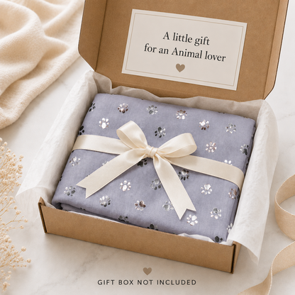 A folded grey scarf with a foil paw print pattern and a cream ribbon bow, presented inside an open kraft gift box on a marble surface with soft neutral props. A card inside reads, 'A little gift for an Animal lover'.