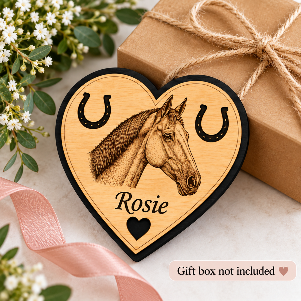 A heart-shaped wooden ornament featuring a engraved horse portrait, horseshoes, and the name 'Rosie', styled on a light surface with flowers, a pink ribbon, and a wrapped gift box in the background. Text at the bottom reads 'Gift box not included'.