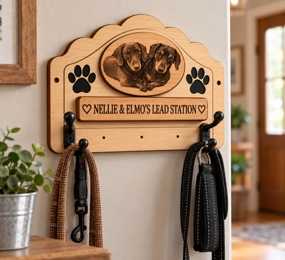 Personalised wooden dog leash rack mounted on a beige wall, featuring a carved portrait of two dachshunds, black paw prints, and the text 'NELLIE & ELMO'S LEAD STATION'. Two black dog leashes hang from the hooks, with a small potted plant on the left and a blurred indoor home background on the right.