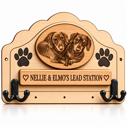 A personalised wooden dog lead rack with two black metal hooks, paw prints, and an oval portrait of two dachshunds. A central plaque reads 'NELLIE & ELMO'S LEAD STATION' with heart symbols.