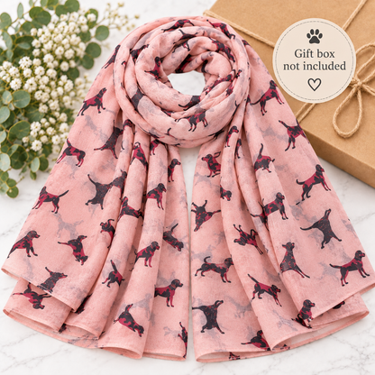 A pink scarf with a repeating pattern of black and brown dogs, styled on a white marble surface next to white flowers and a brown gift box with a 'Gift box not included' sticker.