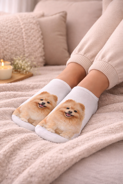 A person sitting on a cosy bed wearing white ankle socks printed with a fluffy cream-colored Pomeranian dog face. The scene is warmly lit with beige bedding, pillows, and a small lit candle in the background.