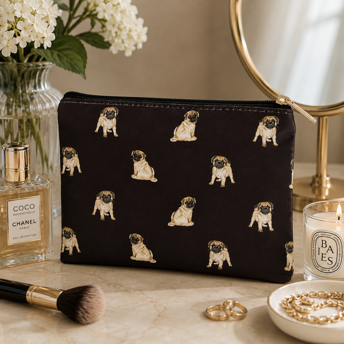 A black zipped cosmetic bag with an all-over print of sitting pug dogs, styled on a marble surface with a Chanel perfume bottle, a mirror, a candle, and jewellery nearby.