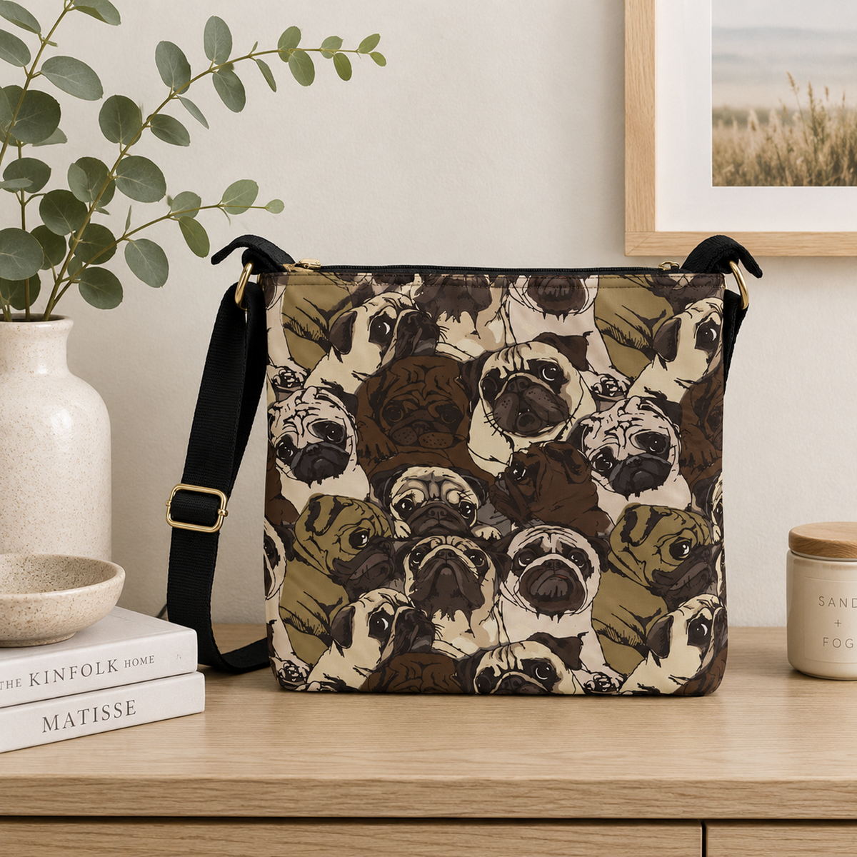 A shoulder bag with a repeating pug dog print pattern, styled on a wooden table next to a vase with greenery and a candle.