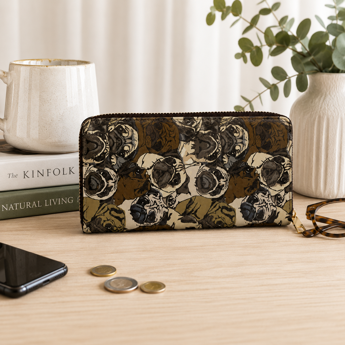 A zip-around wallet with an all-over illustrated pattern of pug faces in olive green, brown, black, and cream, displayed on a light wood tabletop with a mug, books, coins, a phone, and eyeglasses nearby.