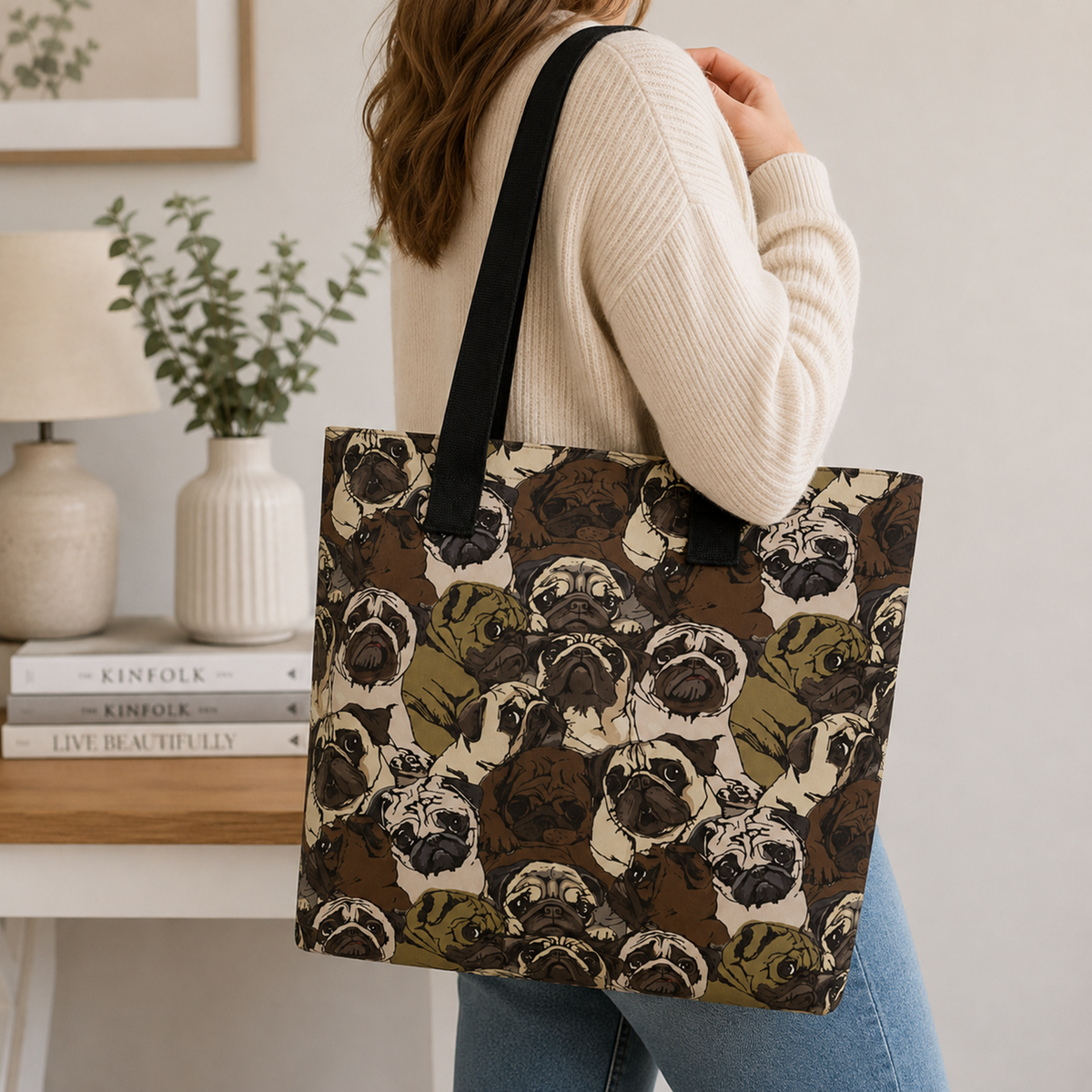 A woman wearing a cream ribbed sweater and blue jeans carries a large tote bag with a black shoulder strap. The tote bag features an all-over illustrated pattern of pug dog faces in brown, beige, and olive green.