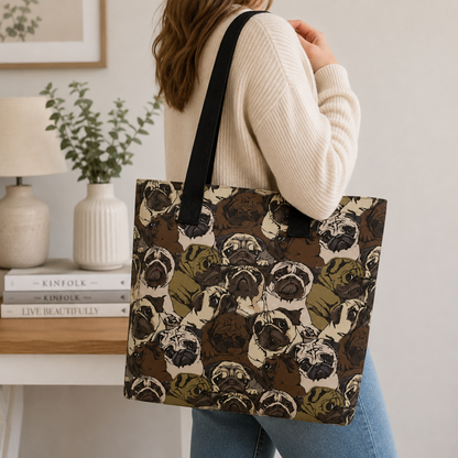 A woman wearing a cream ribbed sweater and blue jeans carries a large tote bag with a black shoulder strap. The tote bag features an all-over illustrated pattern of pug dog faces in brown, beige, and olive green.