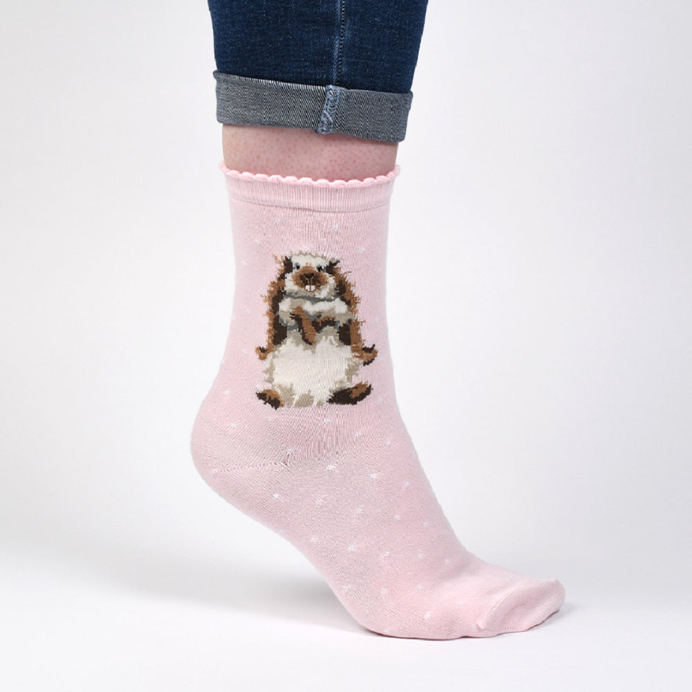 A light pink crew sock with a small hedgehog graphic on the side, worn on a foot with dark blue jeans rolled up, against a plain white studio background.