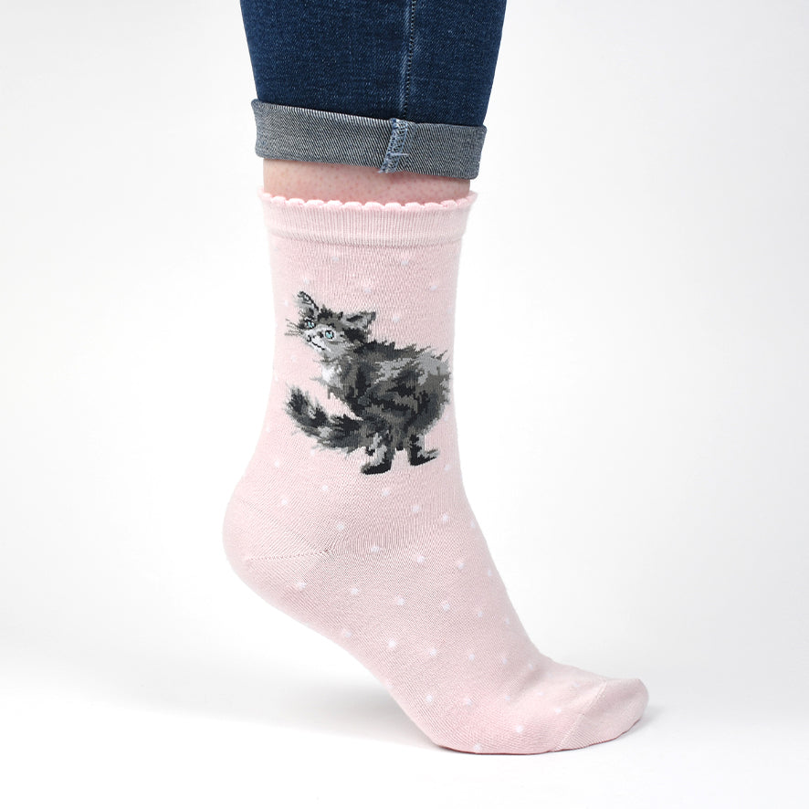 A light pink crew sock with a black and white fluffy cat illustration and small white polka dots, shown on a person's foot with cuffed blue jeans against a white background.