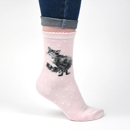 A light pink crew sock with a black and white fluffy cat illustration and small white polka dots, shown on a person's foot with cuffed blue jeans against a white background.