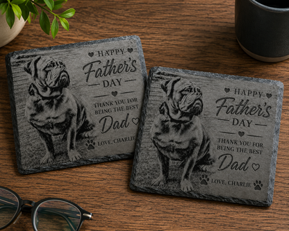 Two personalised slate coasters featuring engraved bulldog illustrations and the text 'Happy Father's Day Thank You for Being the Best Dad Love, Charlie' on a wooden tabletop, with eyeglasses, a plant, and a mug partially visible.