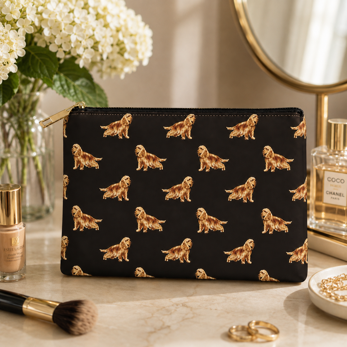 A black zipped cosmetic purse with a repeating pattern of golden Cocker Spaniel dogs in gold, styled on a marble vanity surface with flowers, perfume, and makeup accessories.