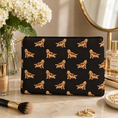 A black zipped cosmetic purse with a repeating pattern of golden Cocker Spaniel dogs in gold, styled on a marble vanity surface with flowers, perfume, and makeup accessories.