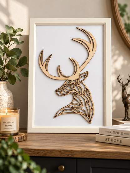 A framed deer wall art piece with a light natural wood stag silhouette on a white background, styled on a wooden cabinet beside a candle, books, and greenery.