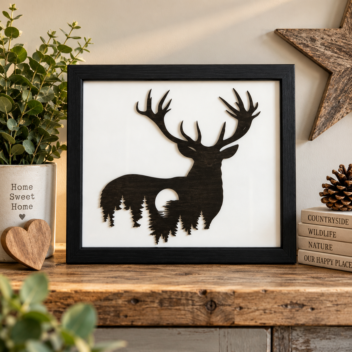 A framed deer silhouette wall art piece with a forest and moon motif inside the deer body, displayed on a rustic wooden shelf with decorative plants, books, and a star ornament.