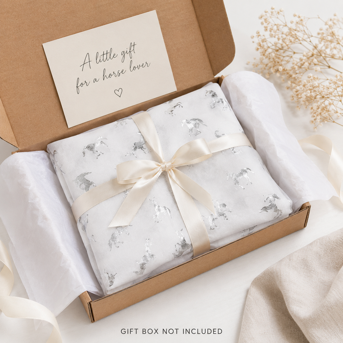 A folded white scarf gift item with a subtle silver unicorn pattern, tied with a cream satin ribbon and placed inside an open kraft gift box. A cream card inside the box reads, 'A little gift for a horse lover.