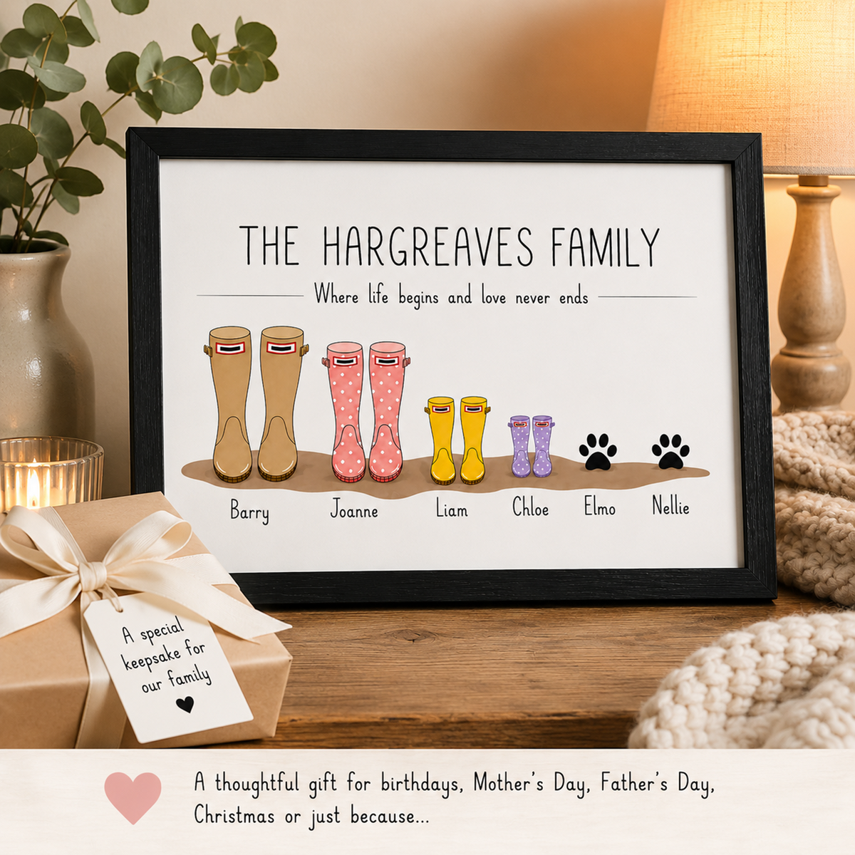 A framed personalised family print featuring the text 'THE HARGREAVES FAMILY' and illustrated rain boots and paw prints with names underneath, displayed on a wooden surface with a gift box and home decor in the background.
