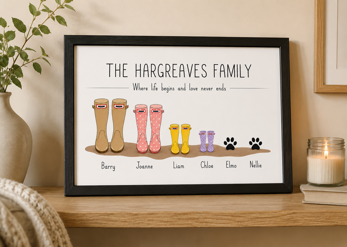 A framed personalised family print featuring the text 'THE HARGREAVES FAMILY' above illustrated rain boots in various colors and sizes, with names labeled beneath each pair. The print is displayed on a wooden shelf next to a vase, a candle, and a book.