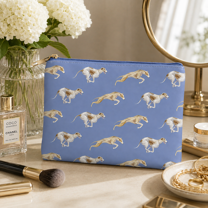 A blue zippered cosmetic bag with a repeating print of greyhounds running, styled on a marble surface with flowers, perfume, makeup brushes, and gold jewellery in the background.