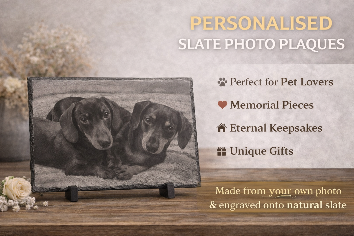 Personalised slate photo plaques displayed on a wooden surface, featuring a black-and-white photo of two dogs engraved on a rectangular natural slate plaque. Text on the right highlights pet memorial keepsakes and custom photo options.