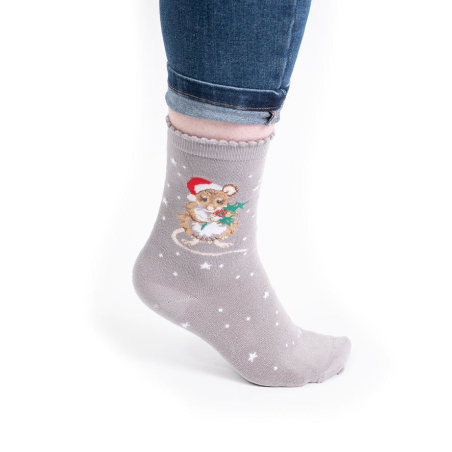 A person's foot wearing a light gray crew sock with a festive graphic of a teddy bear in a Santa hat and scarf, decorated with white stars, paired with blue jeans on a white background.
