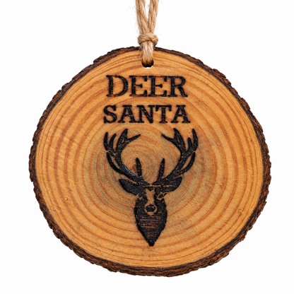 A round wooden Christmas ornament slice with a natural bark edge, featuring a laser-engraved deer head and the words 'DEER SANTA' in black, hanging from a jute twine loop against a plain white background.