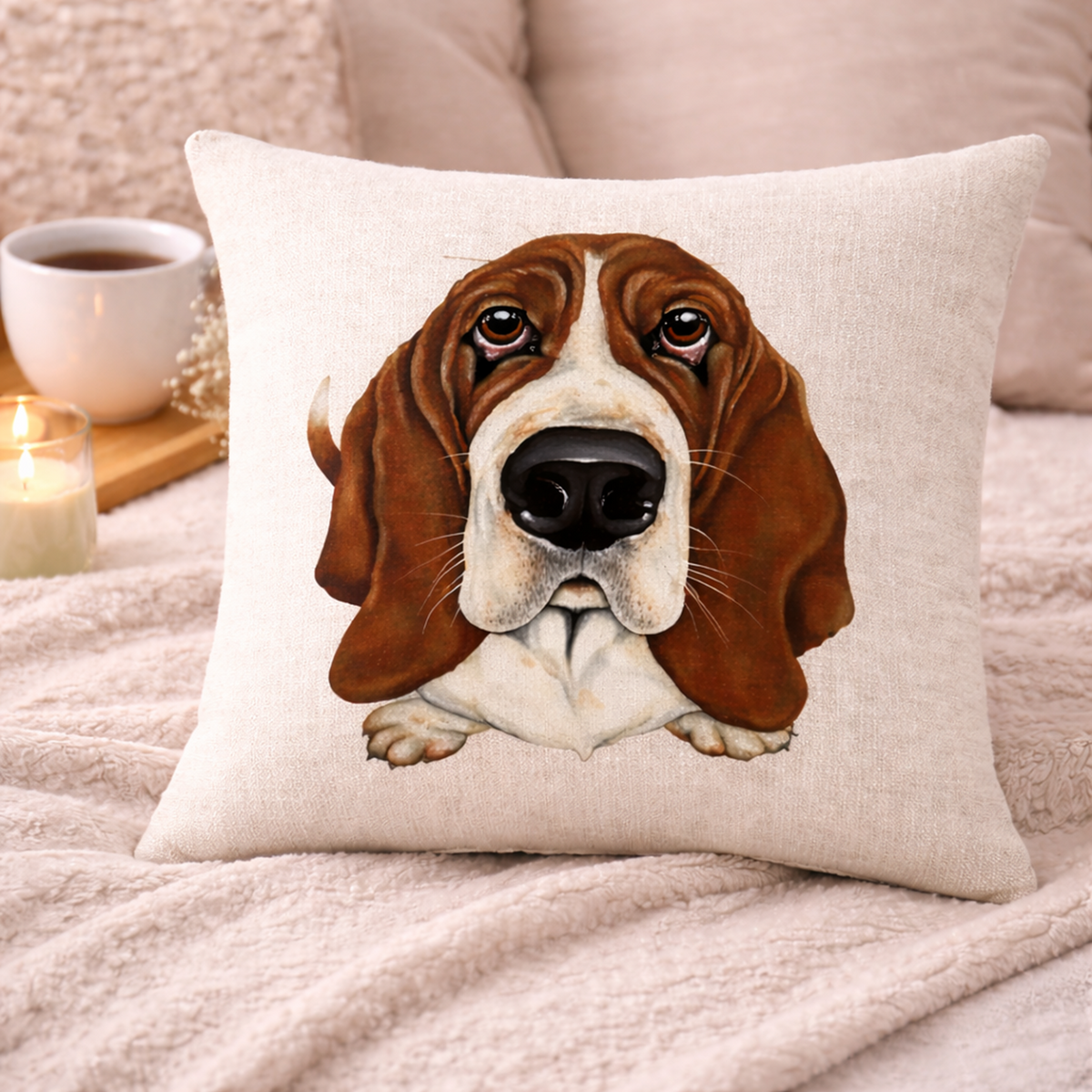 A square cushion cover featuring a printed illustration of a brown and white basset hound, styled on a cosy pink blanket with a cup of coffee and a lit candle in the background.