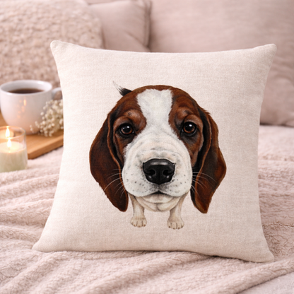 A square beige cushion cover featuring a printed illustration of a brown and white beagle puppy's face, styled on a cosy pink blanket with a mug and candle in the background.