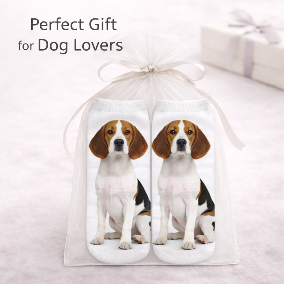 A pair of white trainer socks printed with a seated beagle dog, presented inside a clear tulle gift bag tied with a white ribbon. The image includes the text 'Perfect Gift for Dog Lovers' and features a soft, blurred background with a matching gift box.