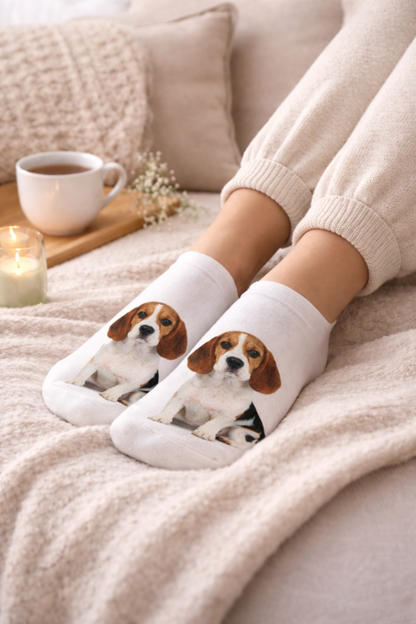 White crew socks with a printed beagle puppy design, worn on a person's feet resting on a cosy beige blanket with a cup of tea and a candle nearby.