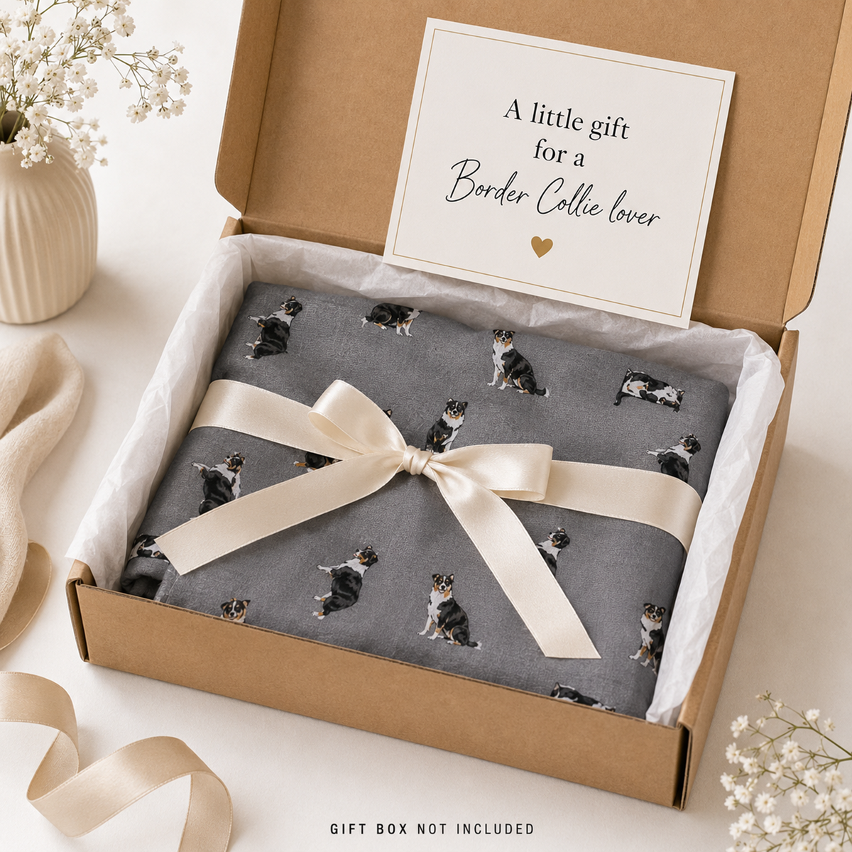 A folded grey scarf gift item printed with tri-coloured Border Collie dogs is tied with a cream satin ribbon inside a brown kraft gift box. An open card inside the box reads, 'A little gift for a Border Collie lover.