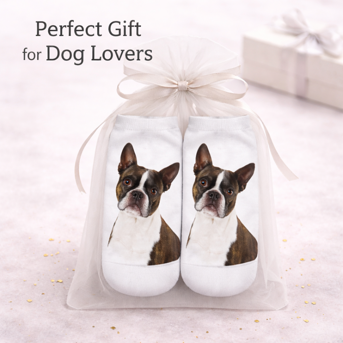 A pair of white socks with printed Boston Terrier dog portraits, presented in a white tulle gift bag tied with a ribbon. The text 'Perfect Gift for Dog Lovers' appears at the top, with a blurred gift box in the background.