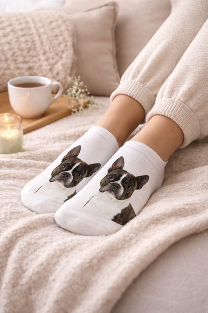 White ankle socks with printed Boston Terrier dog faces, worn by a person sitting on a cozy beige blanket with a cup of coffee, pillow, and candle in the background.