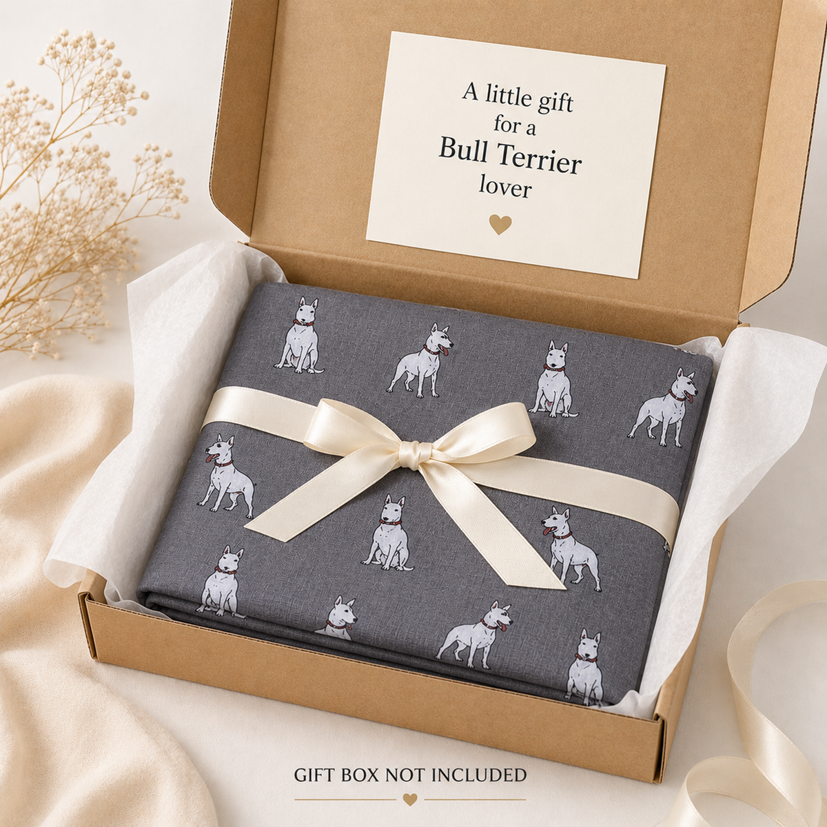 A folded grey scarf item printed with small white Bull Terrier dog illustrations, tied with a cream satin ribbon and presented in a kraft gift box with a card that reads 'A little gift for a Bull Terrier lover.' The scene is styled with soft beige fabric and dried flowers, and text at the bottom says 'GIFT BOX NOT INCLUDED.