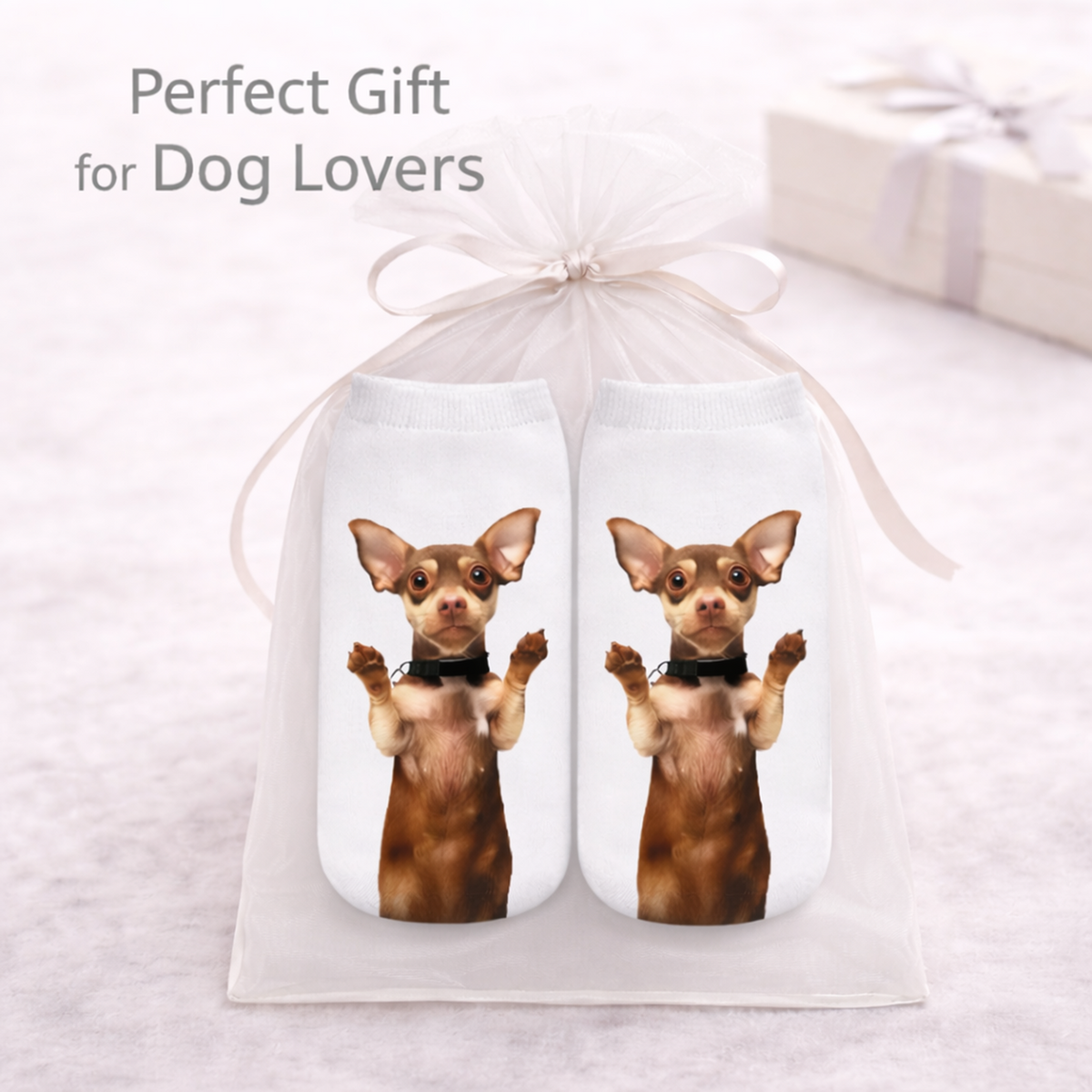 A white sheer gift bag tied with a ribbon, holding two white socks printed with a brown chihuahua dog wearing a black collar. The text 'Perfect Gift for Dog Lovers' appears at the top, with a blurred gift box in the background.
