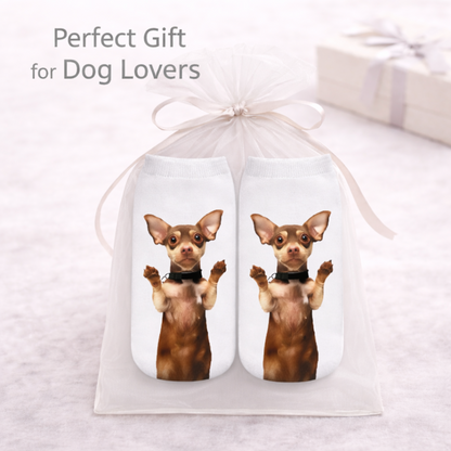 A white sheer gift bag tied with a ribbon, holding two white socks printed with a brown chihuahua dog wearing a black collar. The text 'Perfect Gift for Dog Lovers' appears at the top, with a blurred gift box in the background.