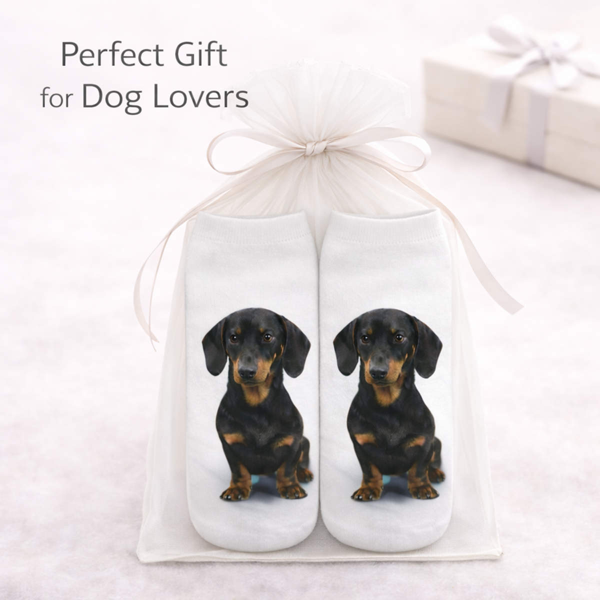A pair of white socks with printed dachshund dog images, presented in a clear gift bag tied with a white ribbon. The text 'Perfect Gift for Dog Lovers' appears in the upper left, with a blurred gift box in the background.