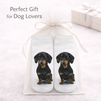 A pair of white socks with printed dachshund dog images, presented in a clear gift bag tied with a white ribbon. The text 'Perfect Gift for Dog Lovers' appears in the upper left, with a blurred gift box in the background.