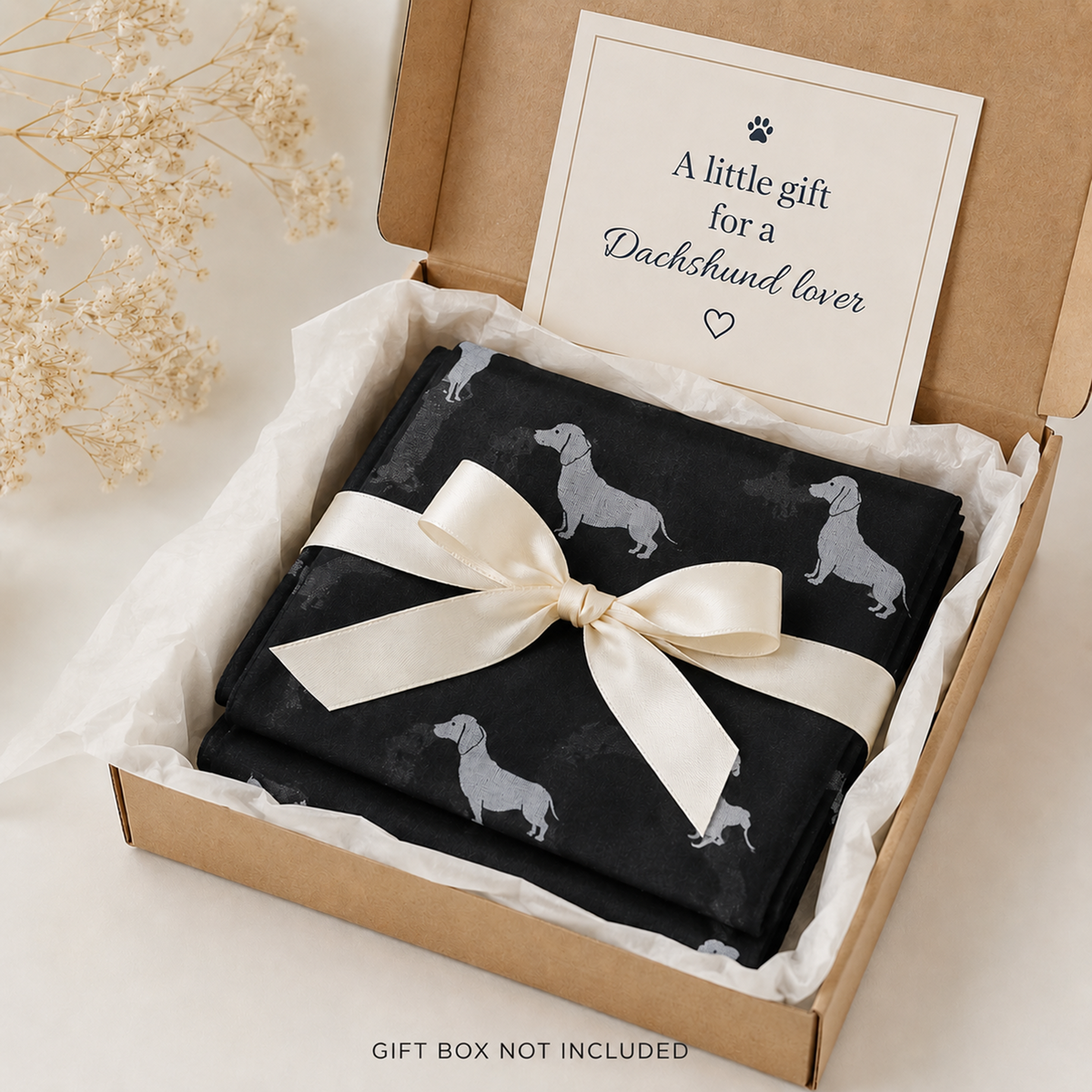 A folded black scarf gift item with a dachshund dog print is presented in a kraft gift box with white tissue paper and a cream satin bow. A printed card inside the box reads, 'A little gift for a Dachshund lover.