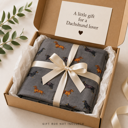 A folded grey scarf gift with a dachshund print, tied with a cream ribbon in a kraft gift box with tissue paper and a card that reads 'A little gift for a Dachshund lover'.