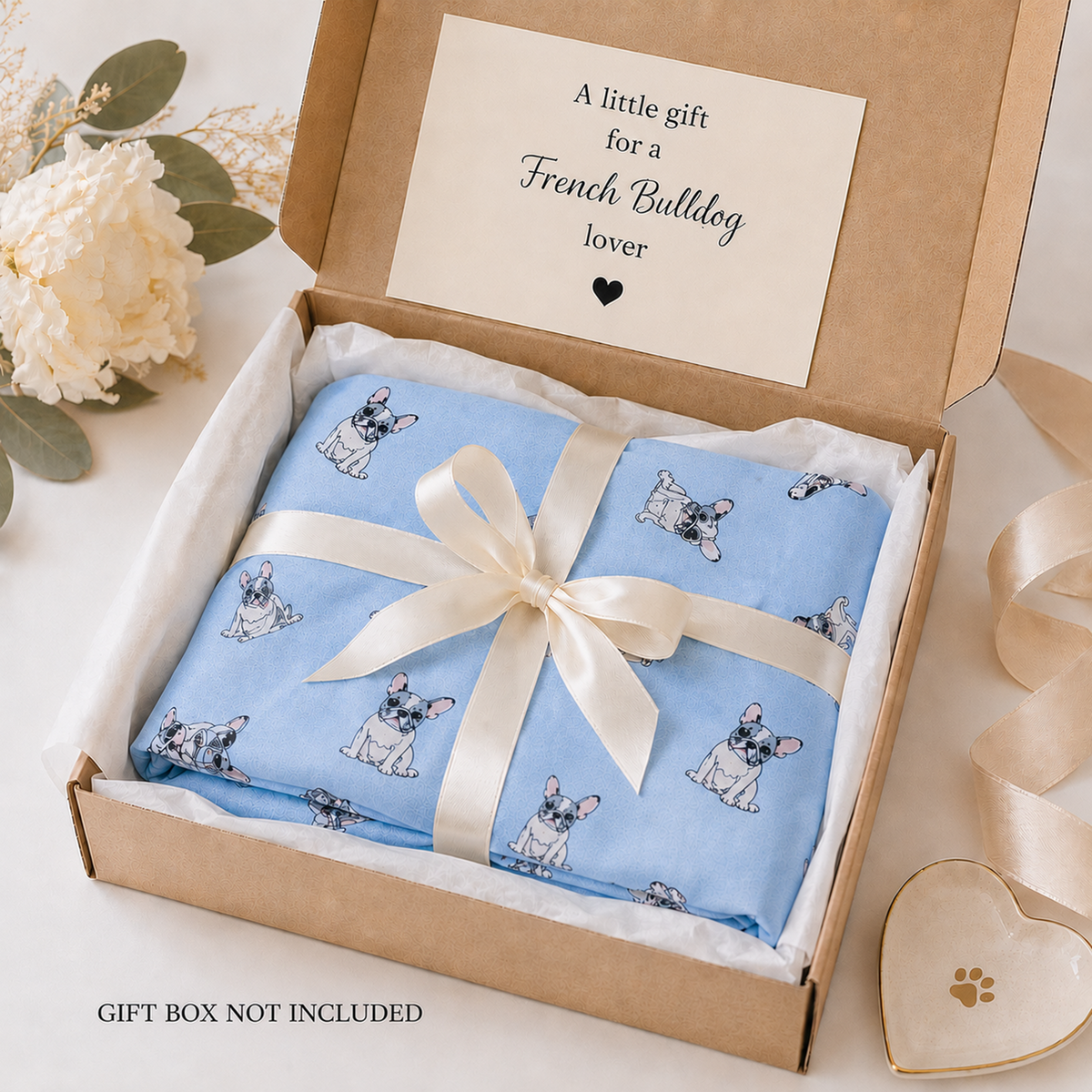 A folded light blue scarf gift item with a French Bulldog print, tied with a cream satin ribbon and presented inside a brown cardboard gift box with a printed message card. White flowers and a heart-shaped dish are styled around the box on a light surface.