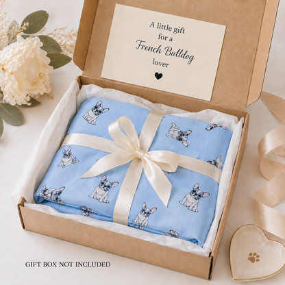 A folded light blue scarf gift item with a French Bulldog print, tied with a cream satin ribbon and presented inside a brown cardboard gift box with a printed message card. White flowers and a heart-shaped dish are styled around the box on a light surface.