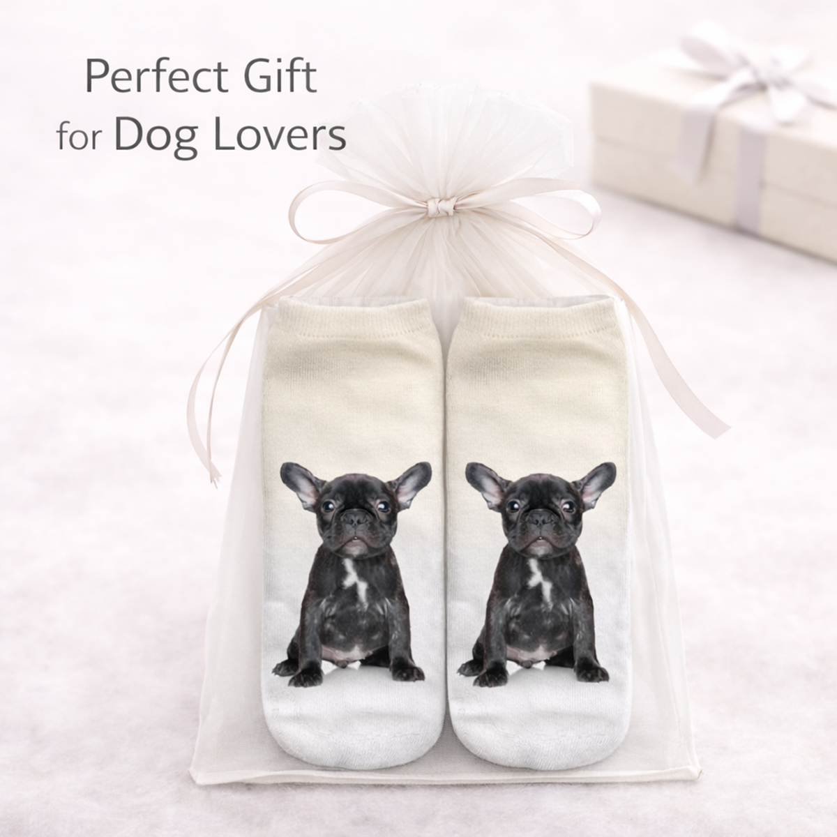A pair of white socks with printed black French bulldog illustrations, presented in a clear gift bag tied with a white ribbon. Text in the upper left reads 'Perfect Gift for Dog Lovers' on a soft light background with a blurred gift box.