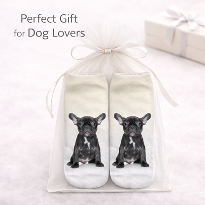 A pair of white socks with printed black French bulldog illustrations, presented in a clear gift bag tied with a white ribbon. Text in the upper left reads 'Perfect Gift for Dog Lovers' on a soft light background with a blurred gift box.