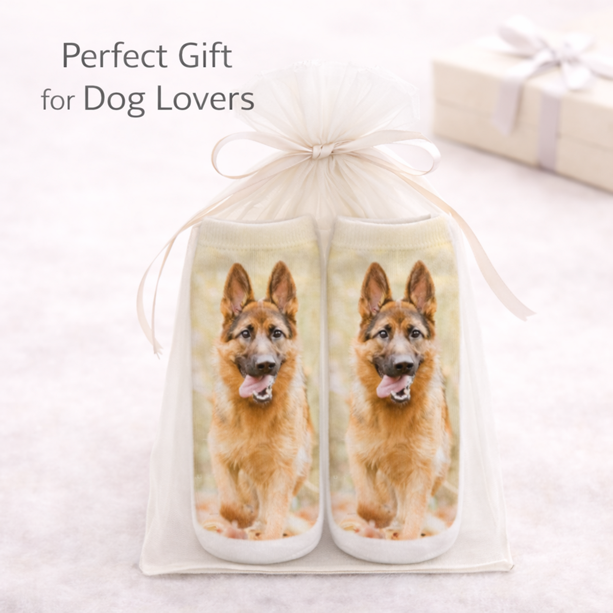 A pair of socks featuring a printed German Shepherd dog, presented in a white tulle gift bag tied with a ribbon. The text 'Perfect Gift for Dog Lovers' is displayed in the upper left, with a blurred gift box in the background.