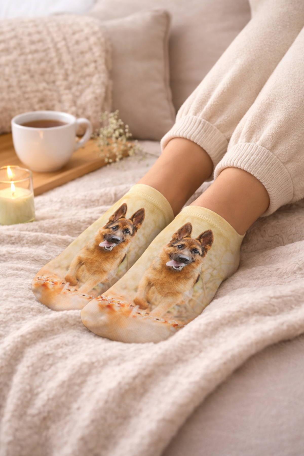 A person sitting on a cozy bed wearing cream-colored socks with a printed image of a German Shepherd dog, with their legs and feet visible in a warm, styled bedroom setting.