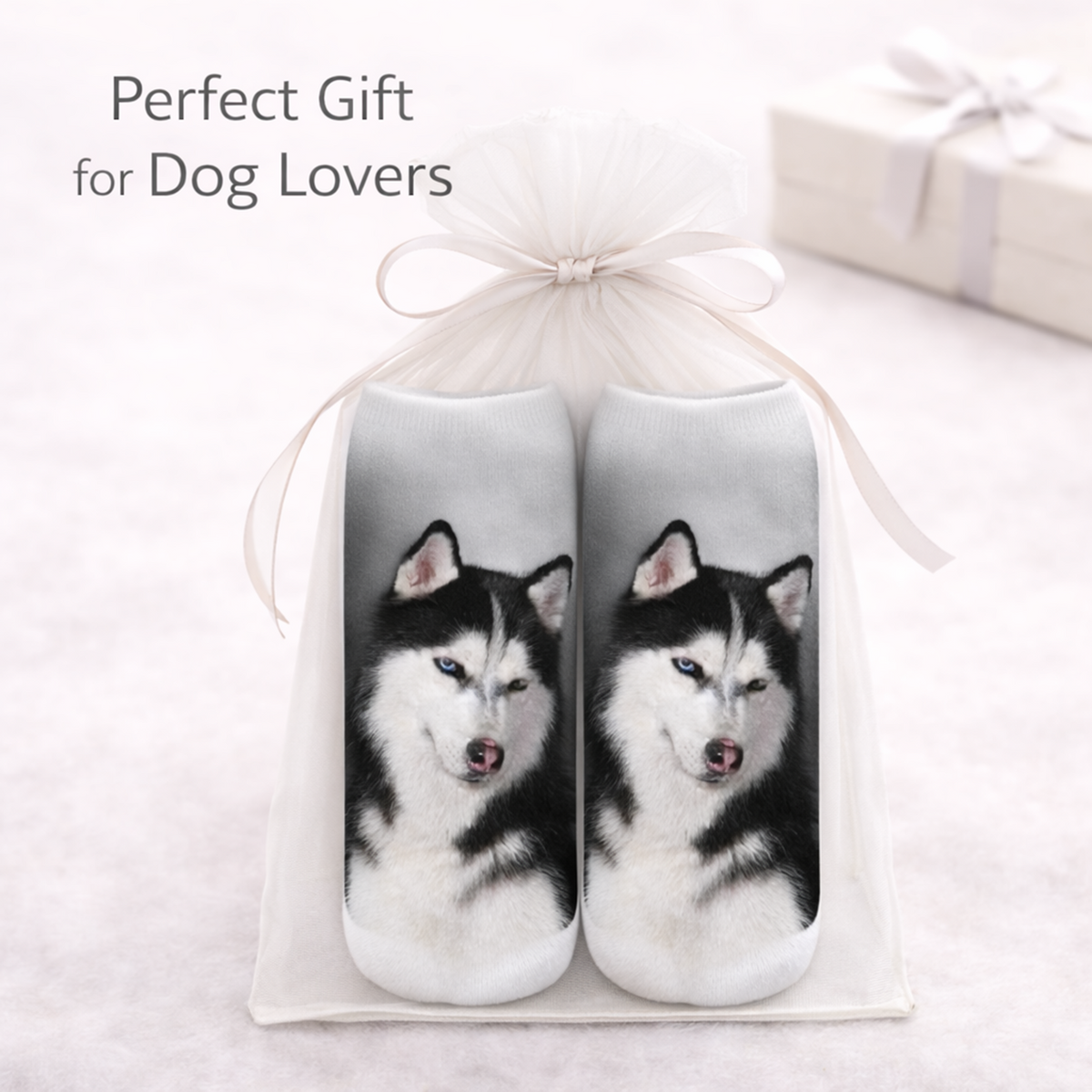 A pair of slip-on socks featuring printed husky dog faces, presented in a white gift bag tied with a ribbon. The text 'Perfect Gift for Dog Lovers' appears in the upper left, with a blurred gift box in the background.