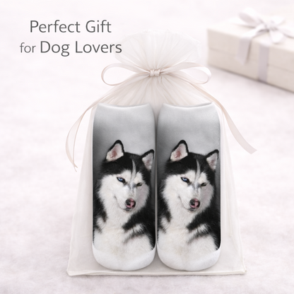 A pair of slip-on socks featuring printed husky dog faces, presented in a white gift bag tied with a ribbon. The text 'Perfect Gift for Dog Lovers' appears in the upper left, with a blurred gift box in the background.